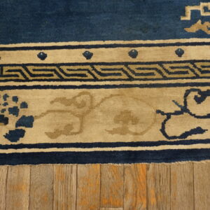 A blue and tan rug with Greek key and floral borders lies on wide distressed wood flooring.