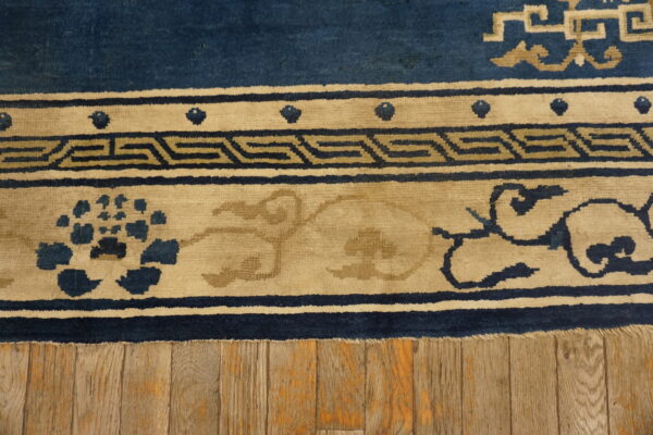A blue and tan rug with greek key and floral borders lies on wide distressed wood flooring.