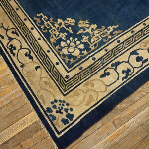 Corner of a low-pile blue rug featuring thick tan geometric and floral borders on aged wooden floorboards.
