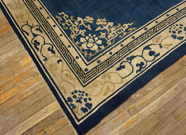 Corner of a low-pile blue rug featuring thick tan geometric and floral borders on aged wooden floorboards.