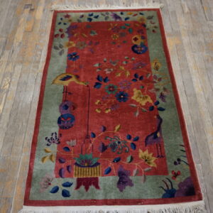 A rectangular rug with a deep red field and light sage border featuring colorful floral and bird patterns lies on distressed wood flooring.