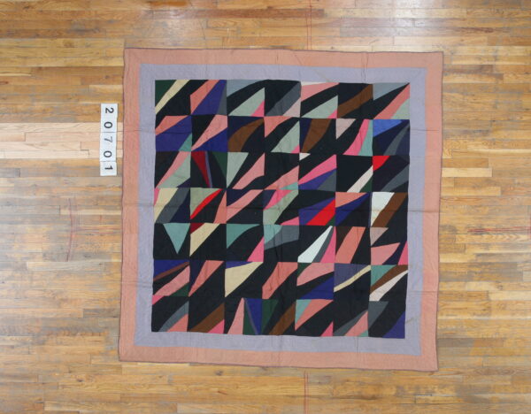 A flat, multicolored geometric textile with mauve and gray borders resting on light plank wood flooring.