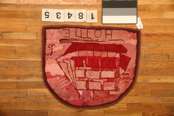Shield-shaped hooked rug in deep red and pink colors with a stylized boat motif on wood.