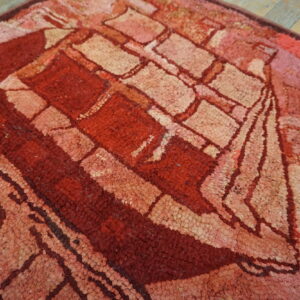 Textured round rug in shades of red and pink with a stylized ship pattern on worn wood flooring.