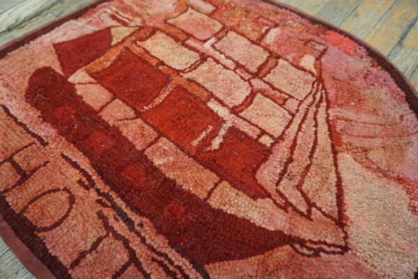 Textured round rug in shades of red and pink with a stylized ship pattern on worn wood flooring.