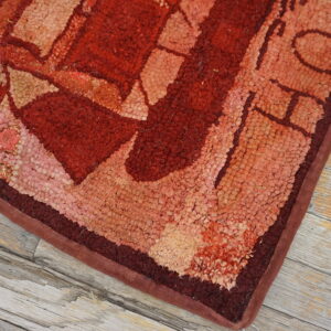 Textured rug corner displaying abstract shapes in reds and tans, resting on distressed gray wood.