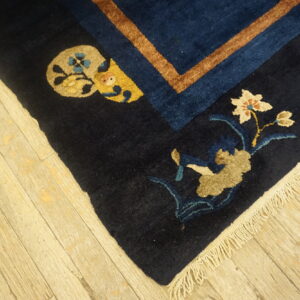 Dark blue rug corner features tan and cream floral motifs, a rust stripe border, and fringe on wood planks.