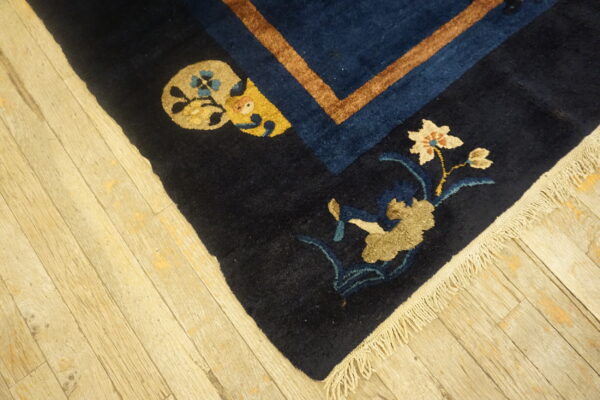 Dark blue rug corner features tan and cream floral motifs, a rust stripe border, and fringe on wood planks.