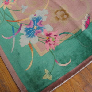 Plush rug with dusty rose field, teal border, and large pink and blue floral motifs on wood flooring.