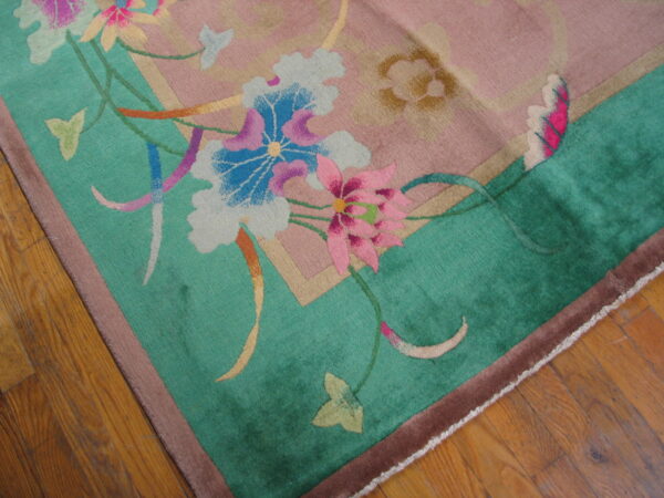 Plush rug with dusty rose field, teal border, and large pink and blue floral motifs on wood flooring.
