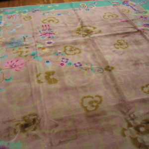 Low-pile rug with a rose field, scrolling patterns, a teal border, and multicolored flowers on wood flooring.