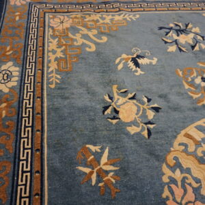 Muted blue rug with tan and cream floral motifs and geometric borders lies on distressed wood floor.