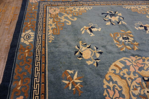 Muted blue rug with tan and cream floral motifs and geometric borders lies on distressed wood floor.