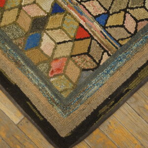 Textured, multicolored hooked rug displaying geometric shapes and thick striped borders resting on distressed hardwood flooring.