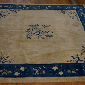 Low-pile square rug featuring a tan field, blue floral corner spandrels, and a wide patterned blue border.