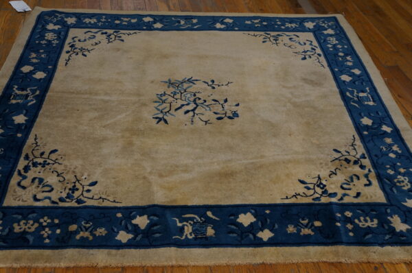 Low-pile square rug featuring a tan field, blue floral corner spandrels, and a wide patterned blue border.