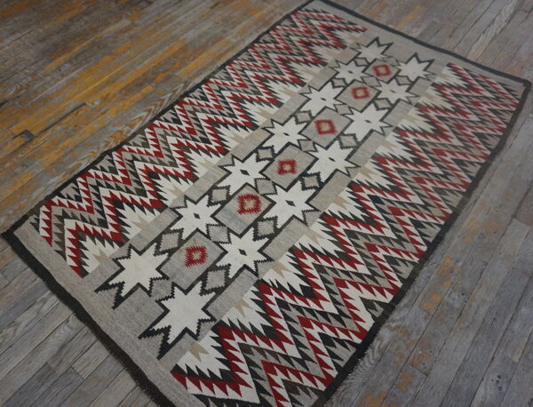 Flatweave rug featuring central geometric stars and zigzag borders in red, gray, cream, and black on wood plank flooring.