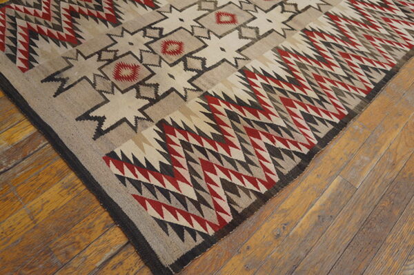 Low-pile geometric rug in beige, red, cream, and dark gray tribal patterns rests on rustic wood flooring.