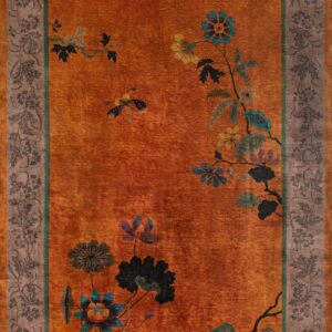 Orange rug with dark blue flowers and a brown patterned border lying on distressed wood flooring.