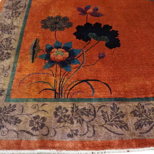 Close-up of a rust-colored rug featuring stylized blue and black floral motifs and a detailed taupe and brown border.