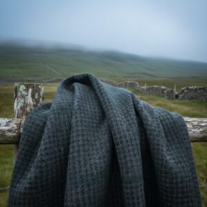 Dark grey houndstooth patterned textile draped over a weathered wooden fence post and rail in a foggy, green landscape – by rugs on net