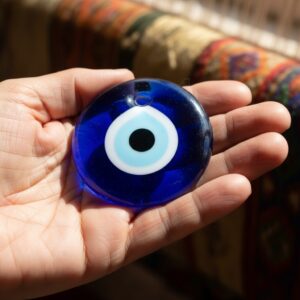 Close-up of a hand holding a blue glass evil eye amulet with a white and light blue center and black pupil – by rugs on net