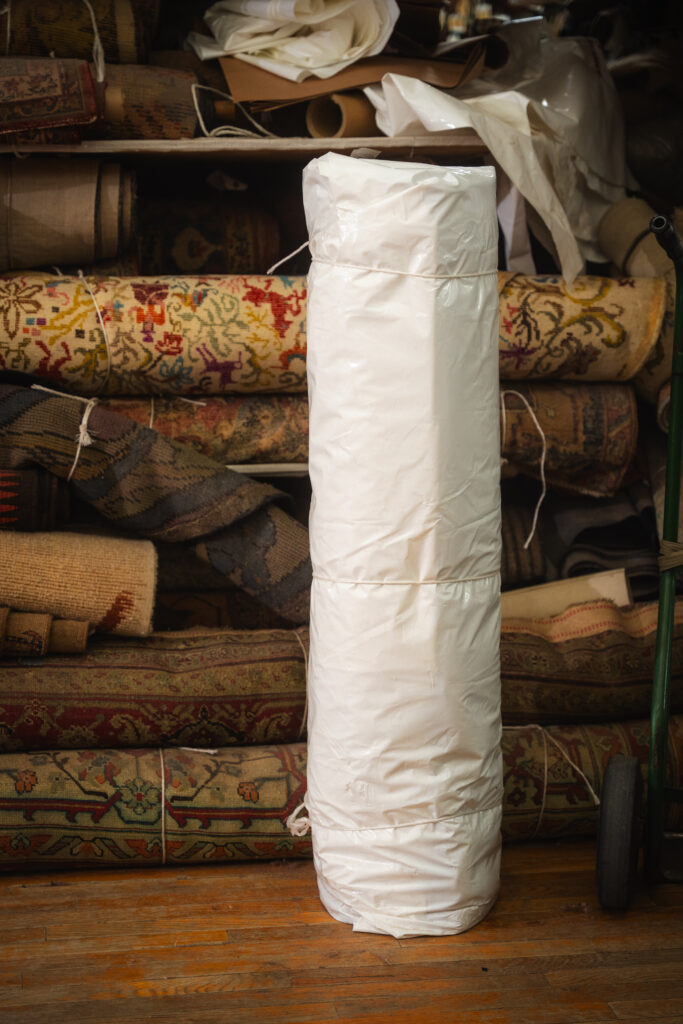 Rolled rug fully wrapped in protective plastic standing in front of stacked rugs