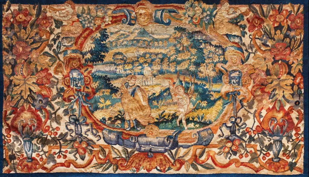 A rectangular, antique flemish tapestry featuring an oval scene of figures in a landscape surrounded by a dense, colorful border of flowers, foliage, and decorative urns, woven in wool and silk threads by rugs on net.