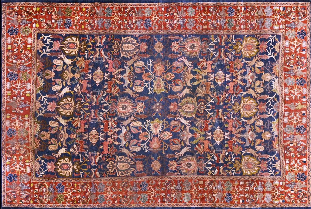 A large, antique persian shah abbas design area rug featuring a dense, allover pattern of stylized flowers and animals in muted earth tones on a dark navy blue field, framed by a wide, rich reddish-orange border by rugs on net.