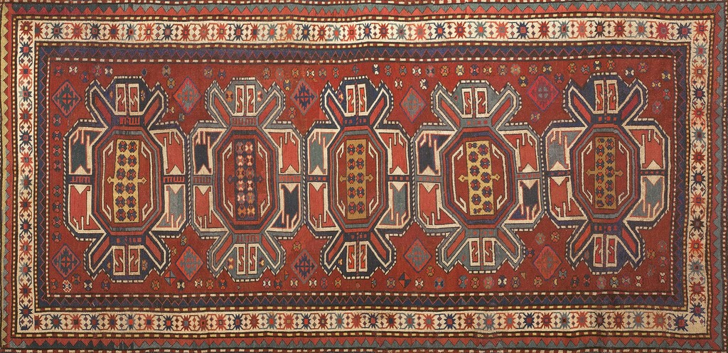 An antique runner-style caucasian rug featuring a repeating pattern of large, angular, geometric medallions in red, navy, gray, and yellow on a deep coral field, bordered by intricate geometric patterns, by rugs on net.