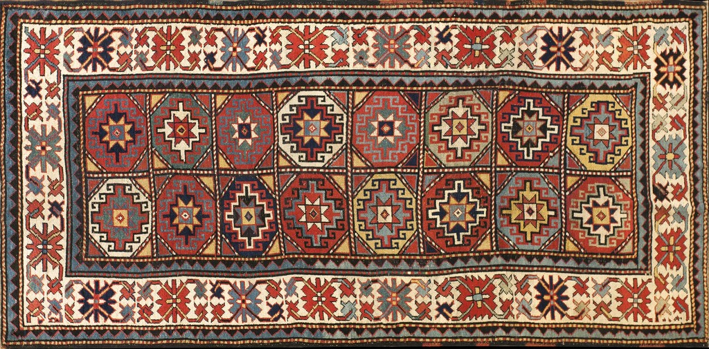 An antique rectangular rug with a repeating grid pattern of stylized octagonal tribal medallions in reds, blues, and yellows, bordered by intricate geometric and floral designs on a cream ground, by rugs on net.