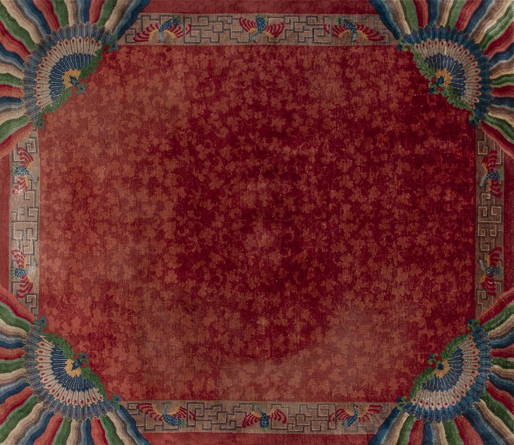 Close-up view of an antique chinese rug with a deep red floral field, geometric border, and prominent corner fan designs in multiple bright colors, by rugs on net.