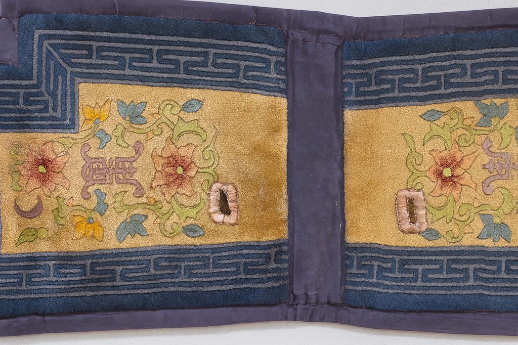 Close-up view of an antique chinese saddle rug showing a gold field with pink and orange floral embroidery bordered by a dark blue greek key pattern and a deep purple binding by rugs on net.