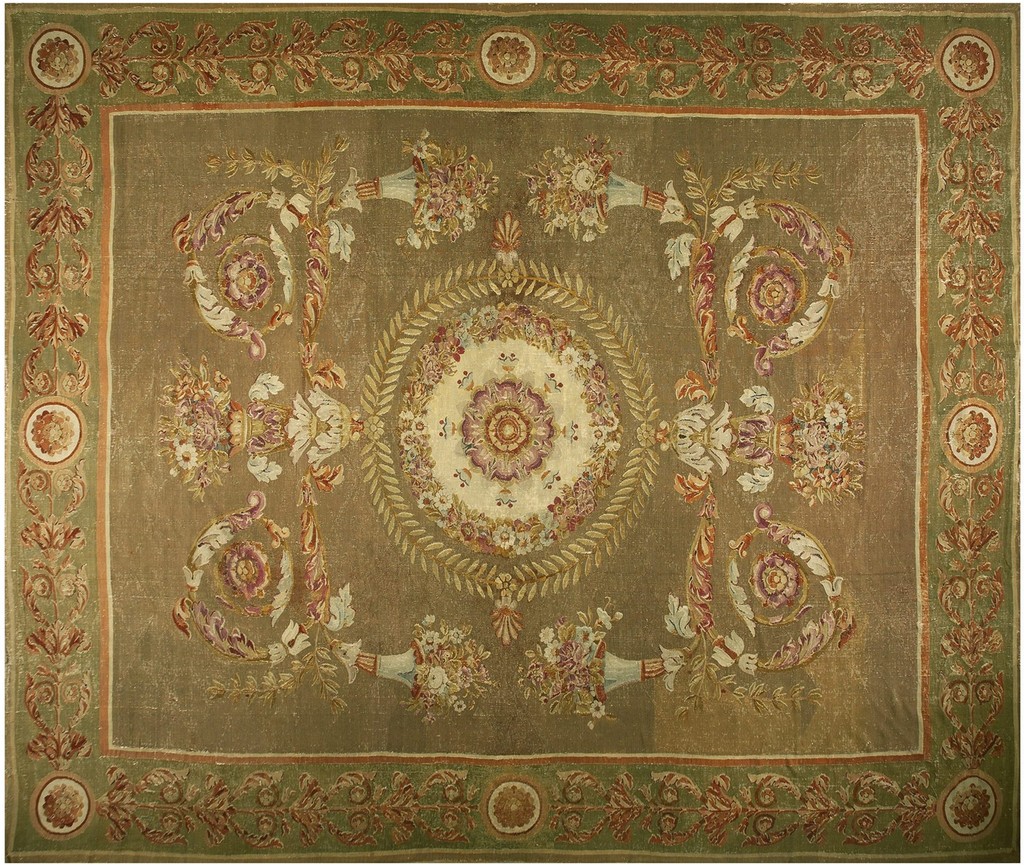A large, antique french aubusson rug with a central cream medallion and a field dominated by olive-brown tones, featuring ornate floral urns and scrolling vine work in pastel colors, framed by a green border by rugs on net.