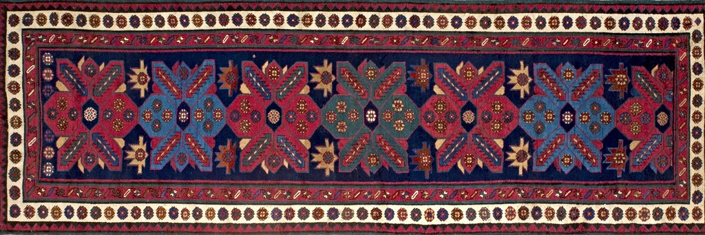 A long, narrow antique caucasian kazak runner rug with repeating large geometric medallions in red, blue, and teal on a dark blue background, framed by detailed borders. By rugs on net