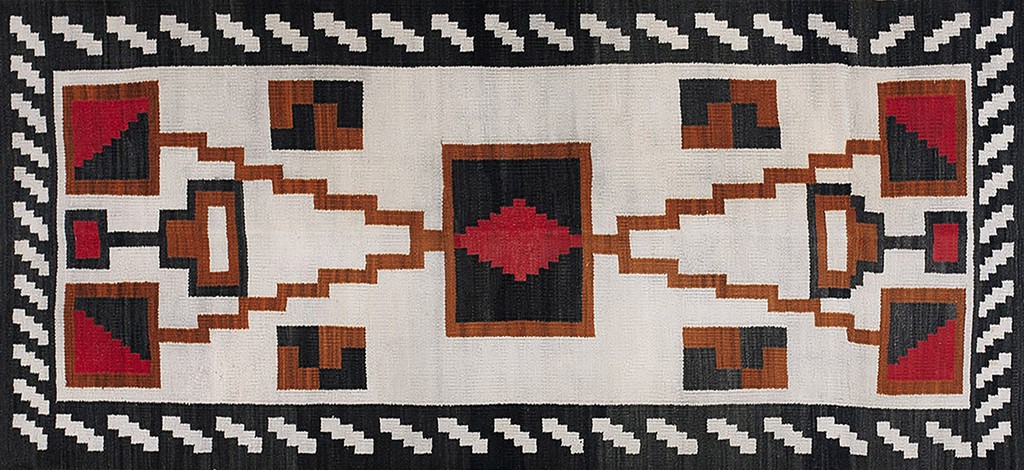 A horizontal antique navajo rug with a bold geometric pattern featuring a central white field contrasted by black, brown, and red rectilinear shapes and a zig-zag border, by rugs on net.