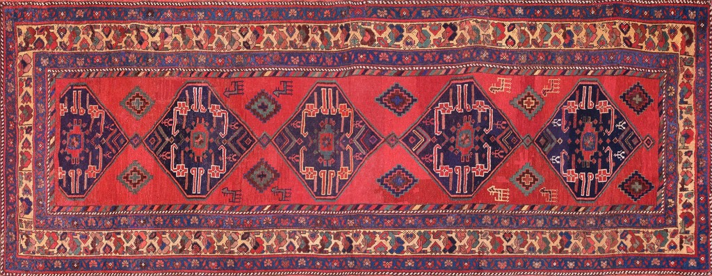An antique persian runner rug featuring a dominant coral-red field with large, repeating indigo diamond medallions filled with tribal patterns, framed by complex geometric and stylized floral borders, by rugs on net.