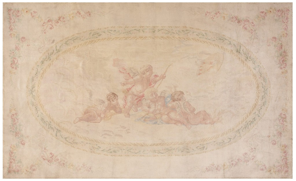 An antique, light-colored rectangular rug featuring a central oval medallion with a painted scene of pink cherubs, bordered by woven garlands and small pink roses on the ivory field, by rugs on net.