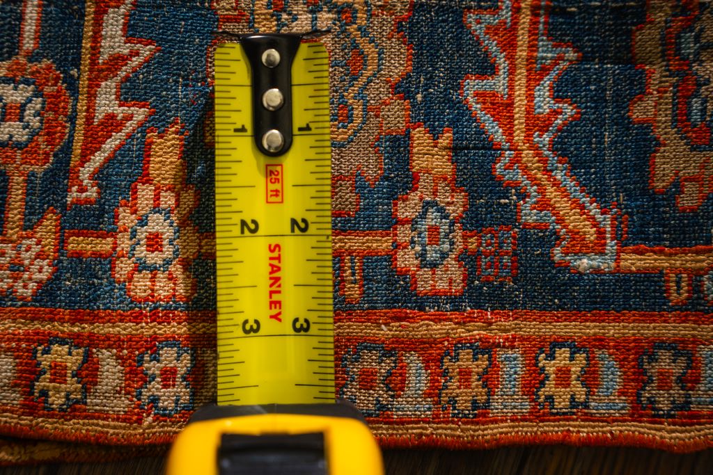 Close-up of a richly colored, traditional rug with a dark blue field and geometric patterns in red and tan, being measured. By rugs on net