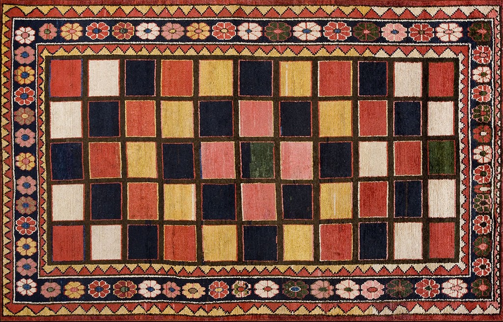 A rectangular antique persian gabbeh rug featuring a central field composed of a grid of multi-colored squares resembling a checkerboard, bordered by a dark indigo band with stylized flowers and geometric patterns. The colors include red, yellow, dark blue, and cream. By rugs on net