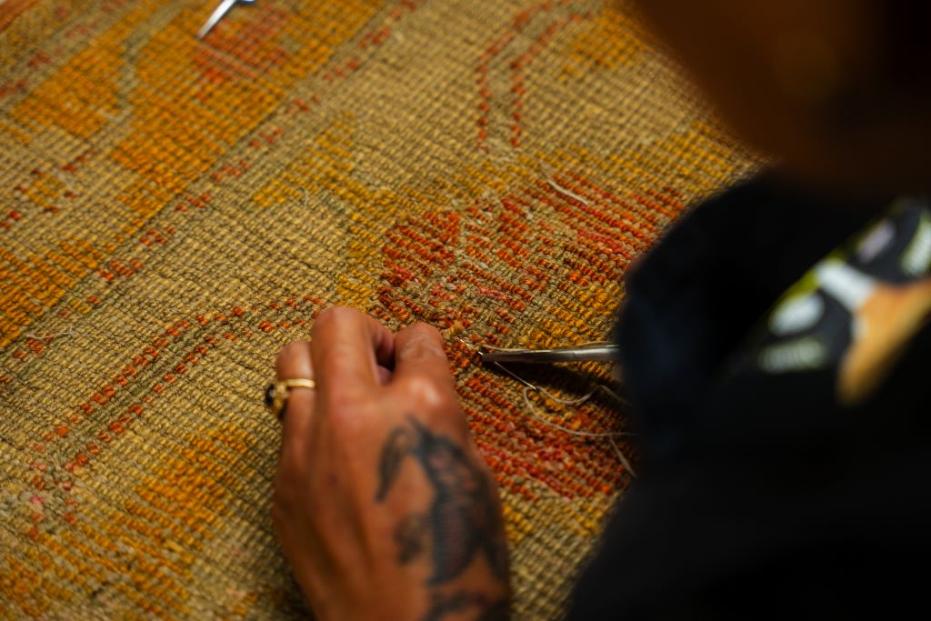 Close-up of a hand with a tattoo carefully sewing or mending a textured, plaid-patterned rug in earthy tones. By rugs on net