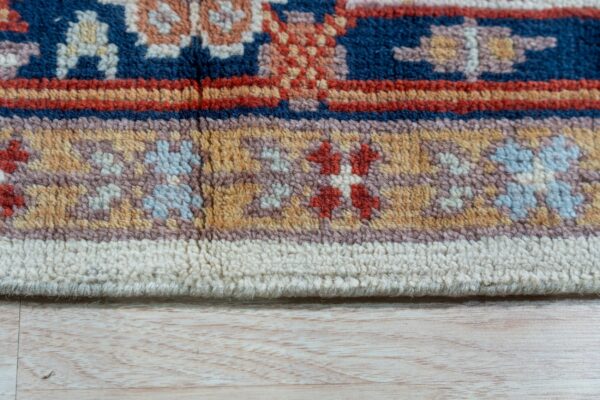 Swatch view of a small red and navy traditional rug on pale wood floors with white furniture and plants.