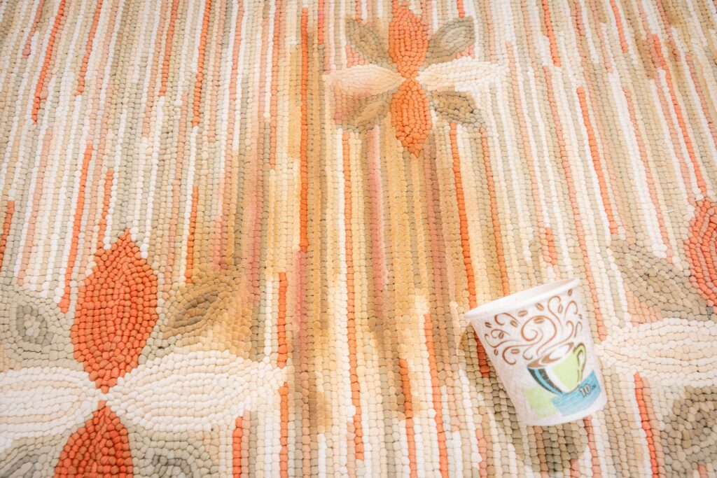 Paper coffee cup tipped over on a handwoven wool rug with warm earth-tone geometric pattern and visible pile texture, by rugs on net