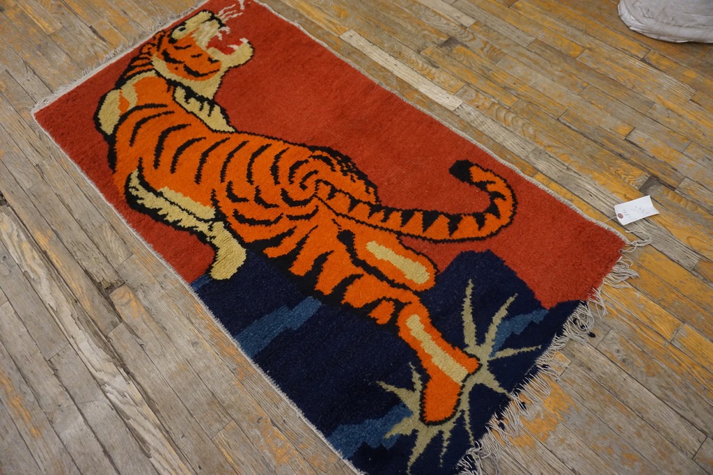 A rectangular rug featuring a large, stylized orange and black tiger roaring against a coral red field, stepping down onto a dark blue, mountainous area, displayed on a rustic wooden floor by rugs on net.