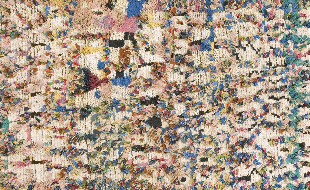 Close-up texture shot of a vibrant, highly textured boucherouitte rug with a cream base dotted heavily with multicolored fabric scraps, including blues, pinks, and black geometric shapes, by rugs on net.
