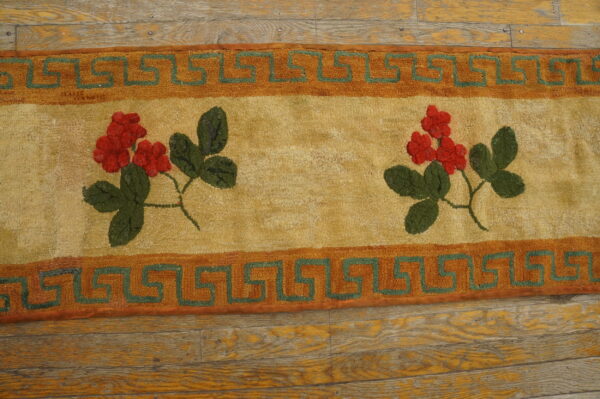 Close-up of a hooked rug featuring red floral clusters and green leaves on a tan field, bordered by an orange and green greek key pattern, by rugs on net