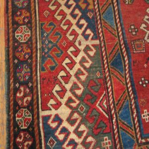 Close-up of a vibrant geometric pattern on a rug, featuring red, blue, and cream colors, next to wooden flooring, by Rugs On Net