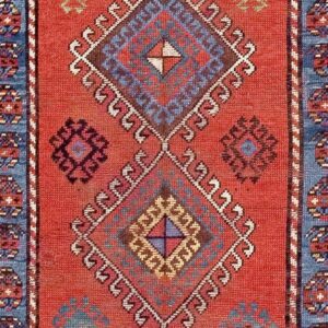 Close-up of a red rug with repeating geometric diamond motifs in blue and brown, bordered by white hooks, by Rugs On Net