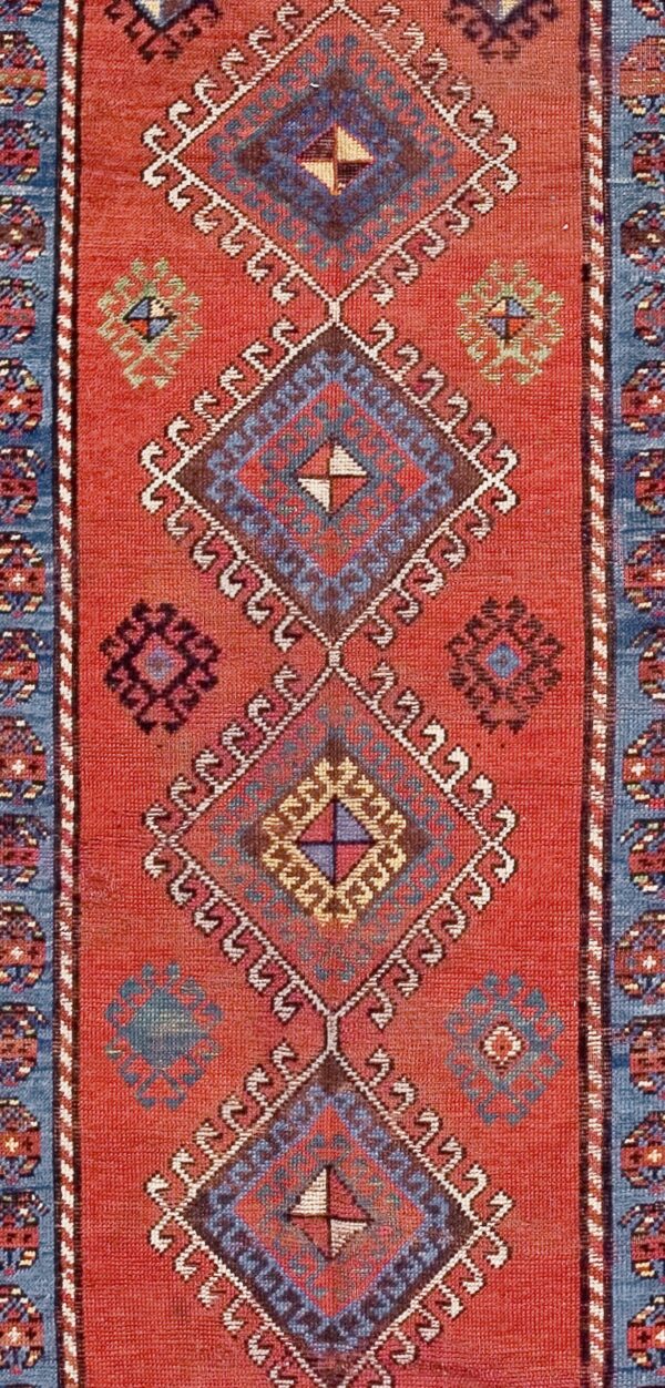 Close-up of a red rug with repeating geometric diamond motifs in blue and brown, bordered by white hooks, by rugs on net
