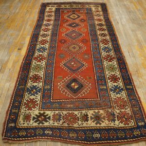 A long runner rug with a dominant red field and geometric diamond motifs, bordered in blue and cream, displayed on a distressed wooden floor by Rugs On Net. by Rugs On Net